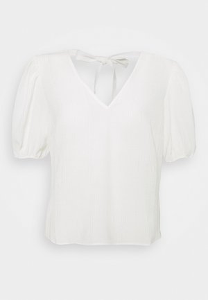 Blouse blanche à manches courtes avec col en V, tissu texturé, manches bouffantes et fermeture par nœud noué au niveau de l’encolure arrière.