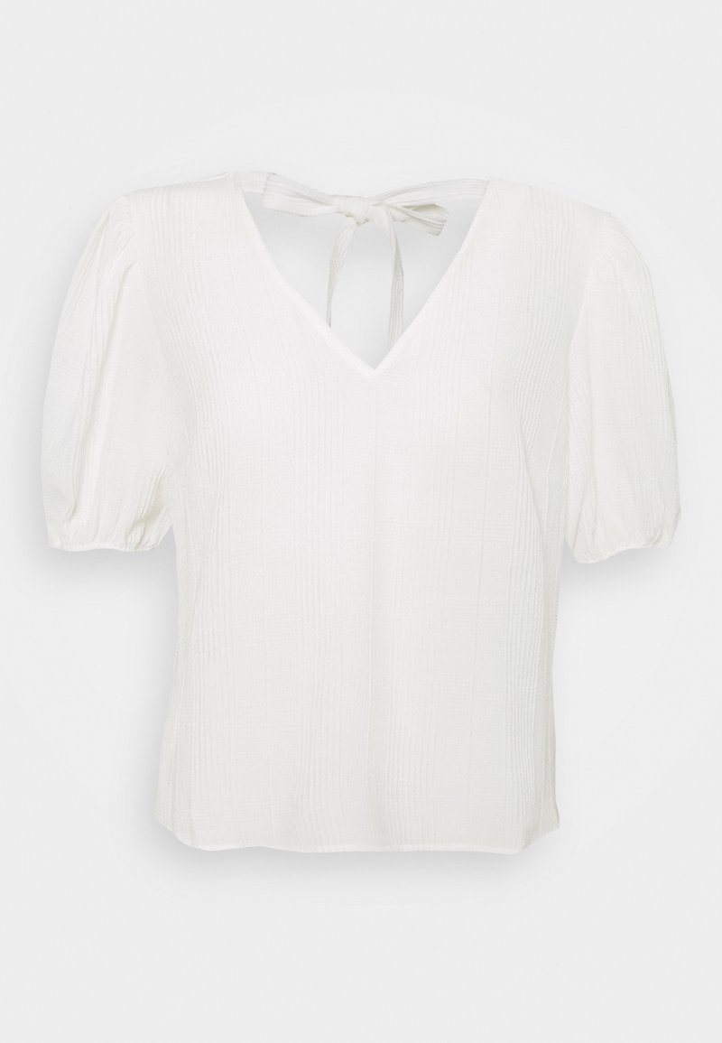 Blouse blanche à manches courtes avec col en V, tissu texturé, manches bouffantes et fermeture par nœud noué au niveau de l’encolure arrière.