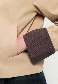 Μπεζ μπουφάν με ομαλή υφή, με μανσέτα σε σκούρο καφέ ριμπ ύφανση και κρυφό τσέπη στο πλάι.