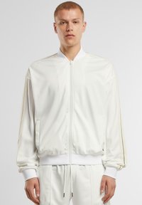 Giacca bomber atletica bianca con chiusura zip, tasche laterali, polsini a coste e strisce in contrasto lungo le maniche. Tessuto liscio e leggero.