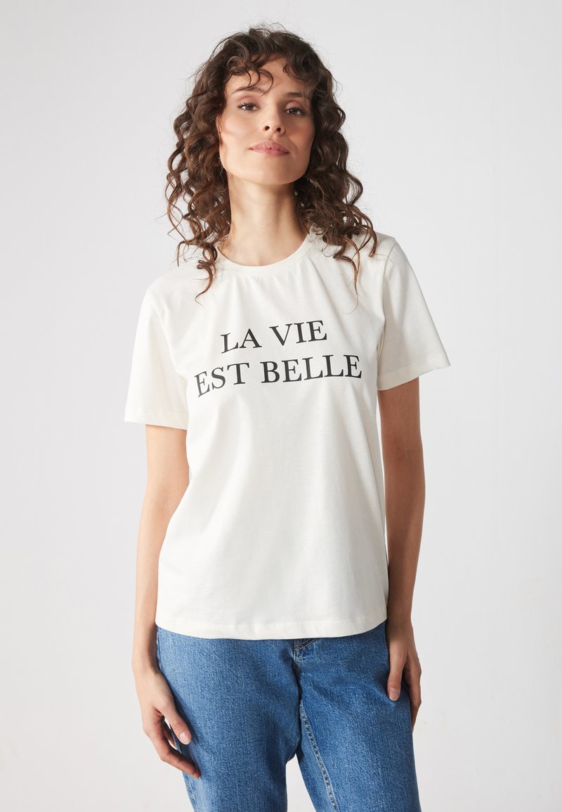 Camisa de algodón blanca con el texto "LA VIE EST BELLE" en letras negras en negrita. Mangas cortas y corte relajado. Combinada con pantalones vaqueros azules.