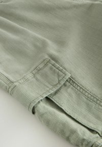 Poignet de manche en tissu vert clair avec des surpiqûres visibles et un détail de patte de fixation sur un fond uni.