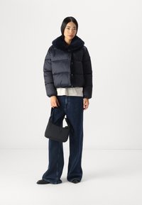 Zwarte pufferjack met een pluche kraag, gecombineerd met een grijze top en brede denim jeans. Model houdt een kleine zwarte tas vast. Zwarte schoenen.
