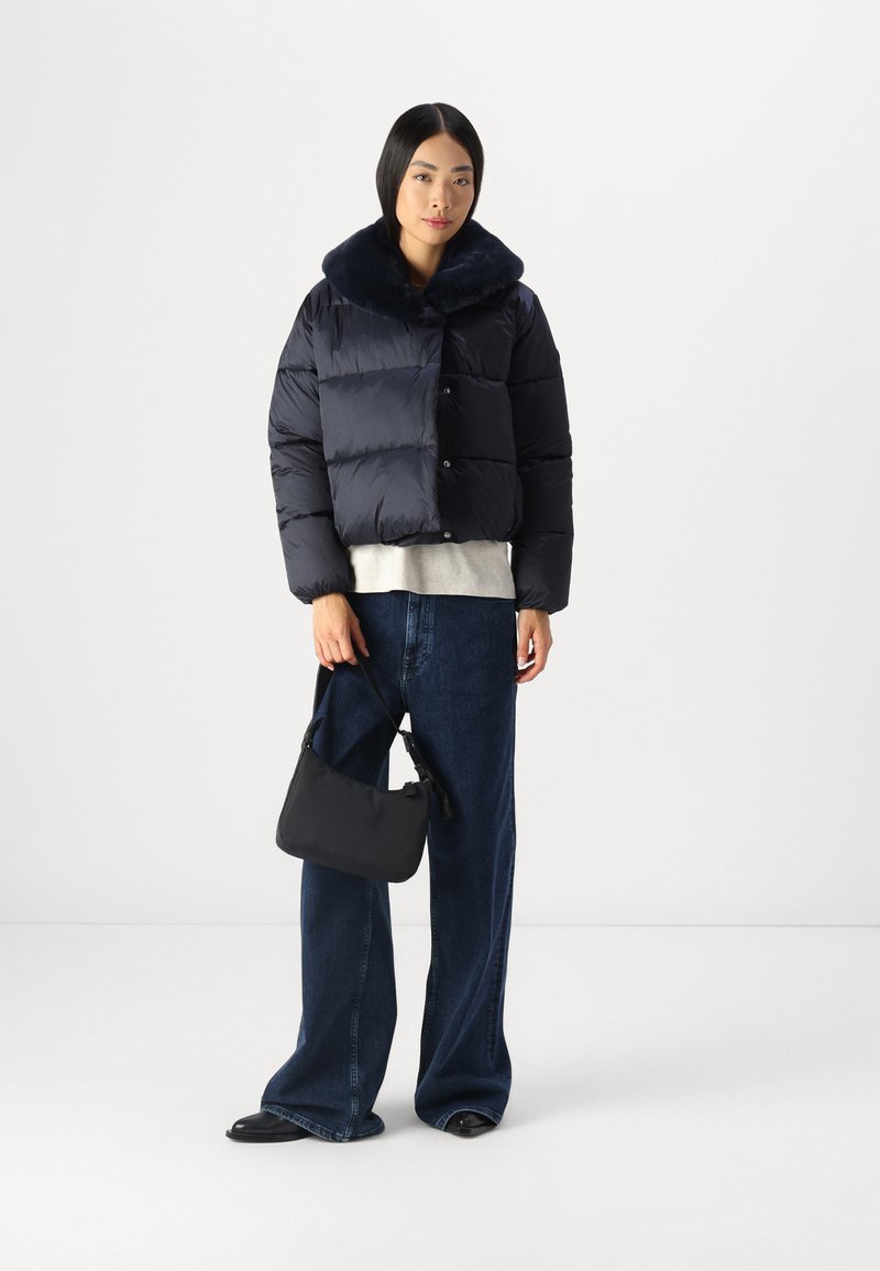 Czarna puchowa kurtka z miękkim kołnierzem, w zestawieniu z szarym topem i dżinsami o szerokich nogawkach. Modelka trzyma małą czarną torbę. Czarne buty.