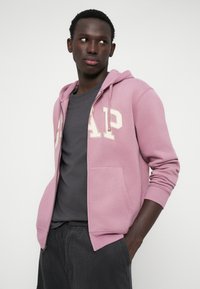 Φόρμα ζακέτα με κουκούλα σε απαλό ροζ χρώμα με κρεμ γράμματα "GAP", μπροστινές τσέπες και ριμπ μανσέτες, συνδυασμένη με σκούρο γκρι t-shirt και παντελόνι.