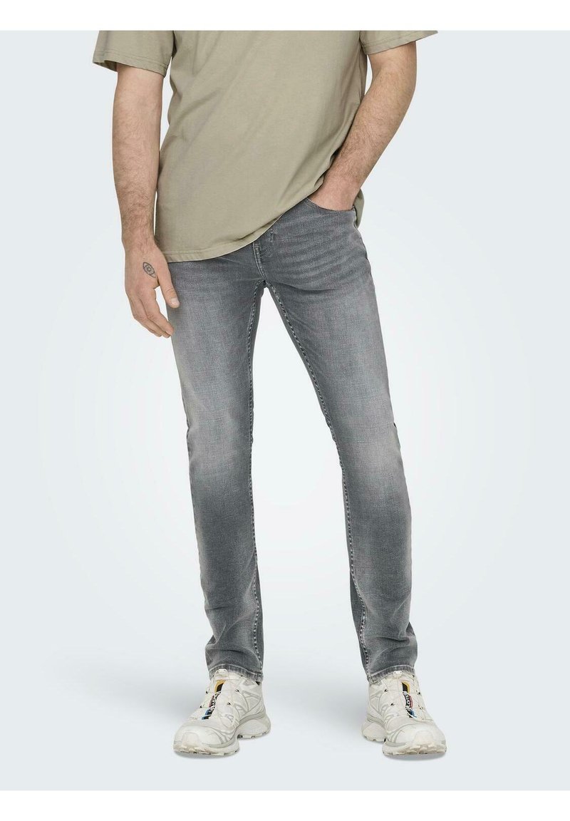Jeans grii slim-fit cu detalii decolorate, purtate cu un tricou lejer de culoare verde măsliniu. Sneakers albi completează aspectul casual.