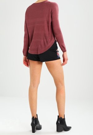Maglione a maglia bordeaux con maniche lunghe, vestibilità comoda e orlo curvo, abbinato a pantaloni corti neri con un motivo decorativo e stivali alla caviglia.