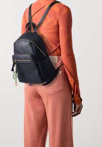 Mochila de cuero negro con cremalleras plateadas, que cuenta con un bolsillo delantero y un charm en forma de cactus. Combinada con pantalones de pierna ancha rosas y una blusa de manga larga.