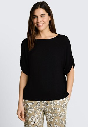 Femme aux longs cheveux bruns, portant un haut noir à manches courtes et un pantalon beige avec des motifs floraux blancs, souriante, les mains dans les poches.