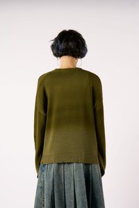 Άτομο με κοντά σκούρα μαλλιά που φορά μεγάλα σκουλαρίκια κρίκους, φανελένιο πράσινο πουλόβερ και πλισέ τζιν φούστα, γυρισμένο μακριά σε λευκό φόντο.