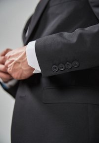 Chaqueta de traje negra con un corte entallado, que presenta un puño de camisa blanca, cuatro botones negros en la manga y una textura de tela suave.