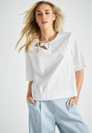 Vrouw met blond haar draagt een witte top met korte mouwen en een zwarte ringdetail bij de halslijn, en lichtblauwe geplooide broek, handen in de zakken.