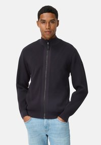 Marineblå jakke med lynlås, høj krave, ribbet tekstur og kontrasterende sømdetaljer. Kombineret med lyseblå denimjeans.