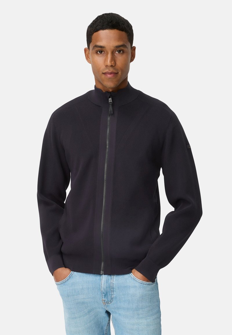 Marineblå jakke med lynlås, høj krave, ribbet tekstur og kontrasterende sømdetaljer. Kombineret med lyseblå denimjeans.