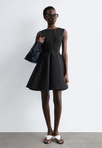 Femme portant une robe noire évasée sans manches, des lunettes de soleil noires, tenant un sac cabas bleu marine et des sandales blanches à enfiler sur un fond uni.