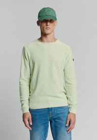 Lichtgroene gebreide trui met een ronde hals en geribde manchetten, gecombineerd met een groene pet. Het model draagt een spijkerbroek en staat neutraal.