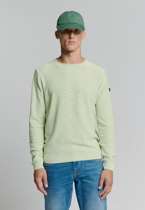 Lichtgroene gebreide trui met een ronde hals en geribde manchetten, gecombineerd met een groene pet. Het model draagt een spijkerbroek en staat neutraal.