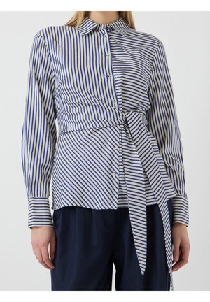 Kvinde iført langærmet skjorte med blå og hvide lodrette striber, bundet i en knude ved taljen, kombineret med mørke bukser.