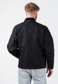 Schwarze Jacke mit einem Cordkragen, die ein schlichtes Design, lange Ärmel und Druckknopfdetails am Saum und an den Ärmelbündchen aufweist.