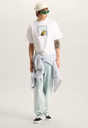 Camiseta blanca de algodón con gráfico de fantasma, vaqueros holgados azul claro, camisa de rayas anudada a la cintura, gafas de sol verdes y zapatillas negras.