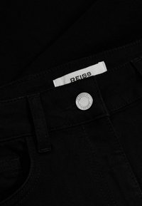 Jean en denim noir au toucher lisse, présentant une fermeture à bouton argenté et une étiquette de marque blanche portant l'inscription "REISS."