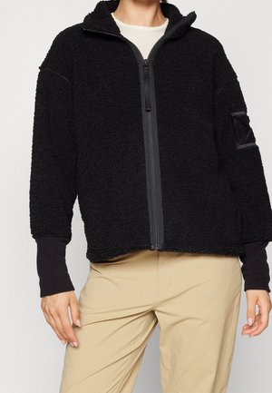 Persoon draagt een zwart fleecejack met ritssluiting over een wit shirt en beige broek, staand met de handen ontspannen langs het lichaam.