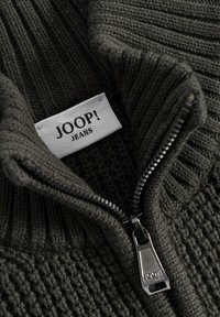 Stickad tröja i mörkgrön med dragkedja, med en ribbad krage och texturerad kropp. "JOOP! JEANS" etikett synlig.