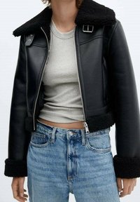 Giacca nera in pelle tagliata con colletto e polsini in pelliccia, caratterizzata da chiusura a zip e trama spessa. Indossata sopra una maglia grigia e jeans blu.