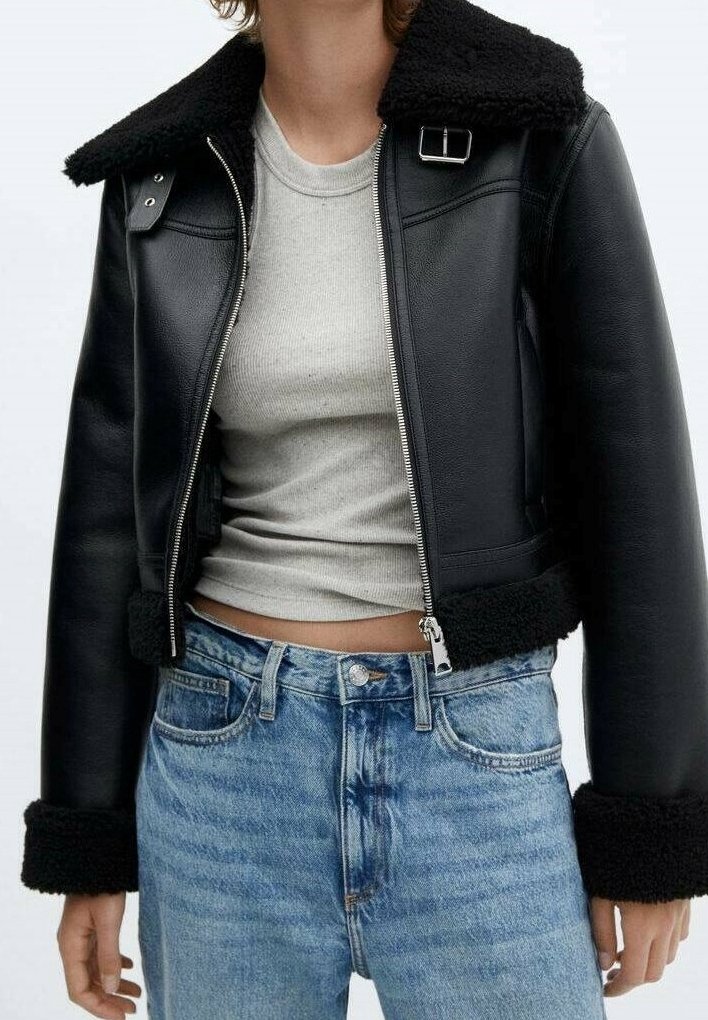 Giacca nera in pelle tagliata con colletto e polsini in pelliccia, caratterizzata da chiusura a zip e trama spessa. Indossata sopra una maglia grigia e jeans blu.
