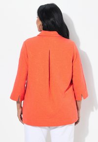 Korálově oranžová košile s 3/4 rukávy, s texturovaným vzorem, záhybem na zadní straně a rozparky na koncích rukávů.