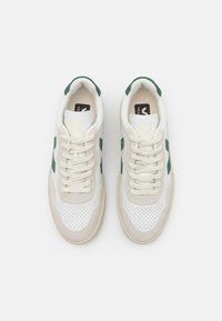 Baskets blanches et beige clair avec des accents verts, présentant un bout perforé, des lacets en tissu et une semelle en caoutchouc texturée.