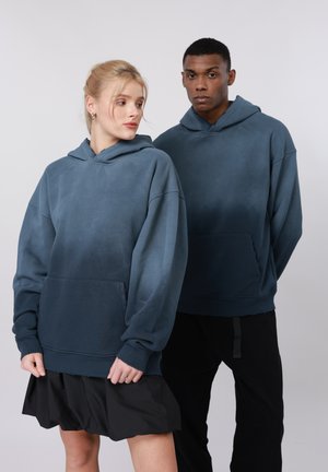 Δύο άτομα που φορούν μπλε φούτερ με κουκούλα· το ένα φοράει μαύρη φούστα, το άλλο μαύρο παντελόνι· ουδέτερο γκρι φόντο.