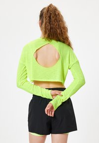 Neon grøn cropped lang�ærmet top med åben rygdetalje, tekstureret stof, parret med sorte shorts, hvilket viser en atletisk stil.
