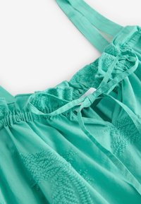 Haut turquoise sans manches avec encolure élastique froncée et détail noué, présentant un motif brodé floral sur tout le tissu.