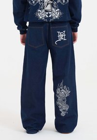 Granatowe dżinsy z dużą białą haftowaną nazwą smoka na lewym nogawce oraz logiem na tylnej kieszeni, o luźnym kroju.