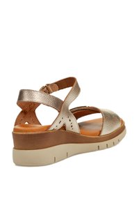 Sandaña metálica dorada con tiras cruzadas, parte superior texturizada, detalles troquelados, plantilla color tostado y suela de cuña en beige claro con líneas de agarre.