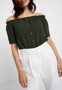 Poisin olkapäiltä pidettävä tummanvihreä paita, jossa on röyhelökaulus, lyhyet hihat ja ruskeat nappikoristeet, yhdistettynä korkeavyötäröisiin valkoisiin housuihin.