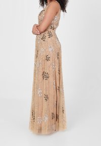 Długa, cielista wieczorowa suknia z przezroczystą warstwą, ozdobiona srebrnymi i czarnymi wzorami kwiatowymi oraz delikatnymi koralikami.