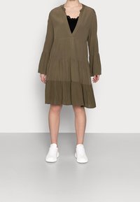 Olivgrön flerlagad klänning med långa ärmar, V-ringning och volangärmar. Matchad med vita sneakers mot en enkel bakgrund.