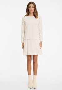 Helles beiges Kleid mit geripptem Muster, langen Ärmeln und einem plissierten Rock. Kombiniert mit hellen, enganliegenden Stiefeletten.