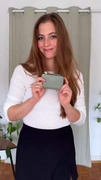 Carteira verde de couro com superfície texturizada, fecho dourado e logotipo da marca. Segurada por uma pessoa vestindo uma blusa branca e saia preta.