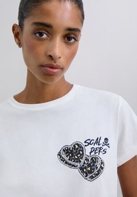 Kobieta z wciągniętymi włosami, ubrana w białą koszulkę z nadrukiem dwóch serc w panterkę oraz napisem "SCAL PERS" z małym symbolem czaszki.