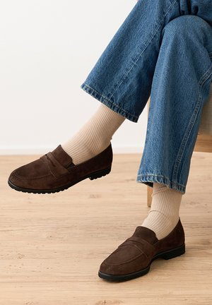 Braune Wildleder-Loafer getragen mit hellbeigen, gerippten Socken und blauen Denim-Jeans, ein Bein über das andere geschlagen, während man auf einem hellen Holzboden sitzt.