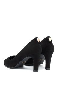 Par de zapatos de tacón alto de ante negro con puntera redondeada, tacones de 7 cm y acentos en forma de logo circular dorado en la parte posterior.