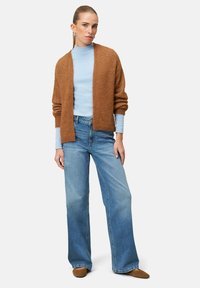 Cardigan marron pelucheux porté sur un col roulé bleu clair, associé à un jean bleu clair à jambes larges et des chaussures plates couleur beige. Design simple et décontracté.