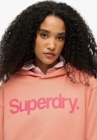Roze katoenen hoodie met een groot "Superdry" logo in een donkerder roze. Draagt over een lichtroze geblokte shirt, met krullend haar zichtbaar.