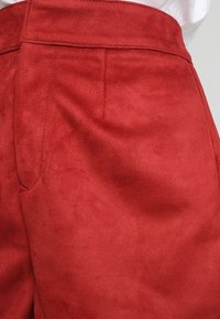 Pantaloni in suede rosso con una texture liscia, caratterizzati da tasche anteriori piatte e una vita senza cuciture. Il colore è ricco e vibrante, arricchendo il design.