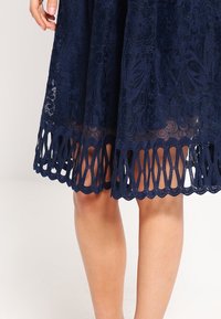 Abito in pizzo blu navy con motivo floreale e orlo a onde. La parte inferiore presenta dettagliati ritagli circolari.