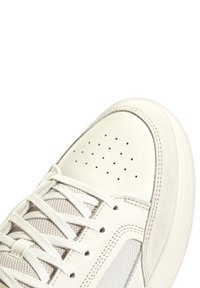 Zapatilla blanca con parte superior de cuero texturizado, puntera perforada, acentos de malla y cordones planos. Suela de goma suave y detalles de costuras mínimos.