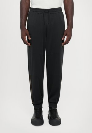 Black athletic pants with elastic waistband, side pockets, and gold accent stripes. Fabric is smooth with a relaxed fit and tapered cuffs.